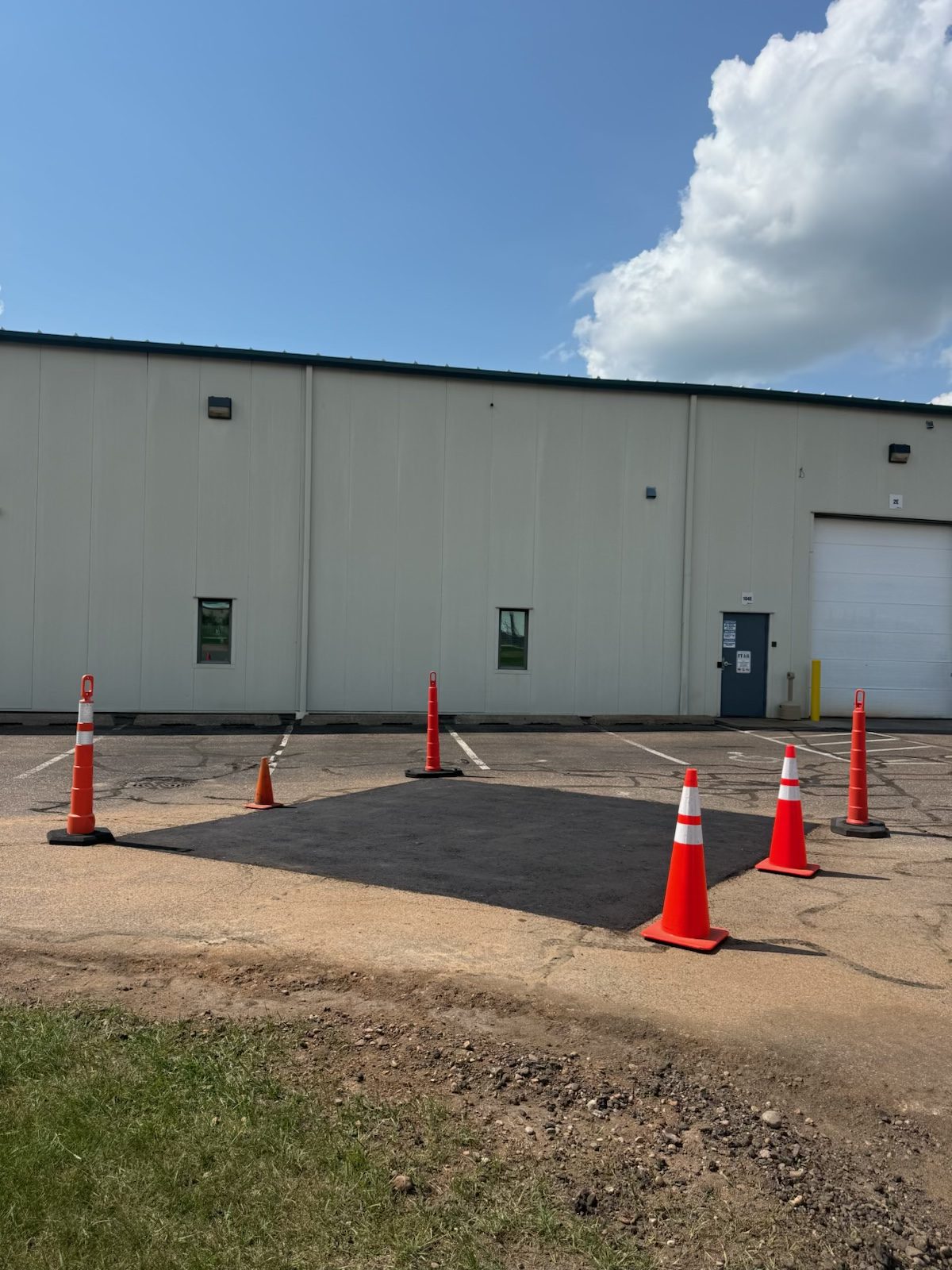 A fresh full-depth asphalt patch coned off in a commercial lot outside an industrial building with dock doors