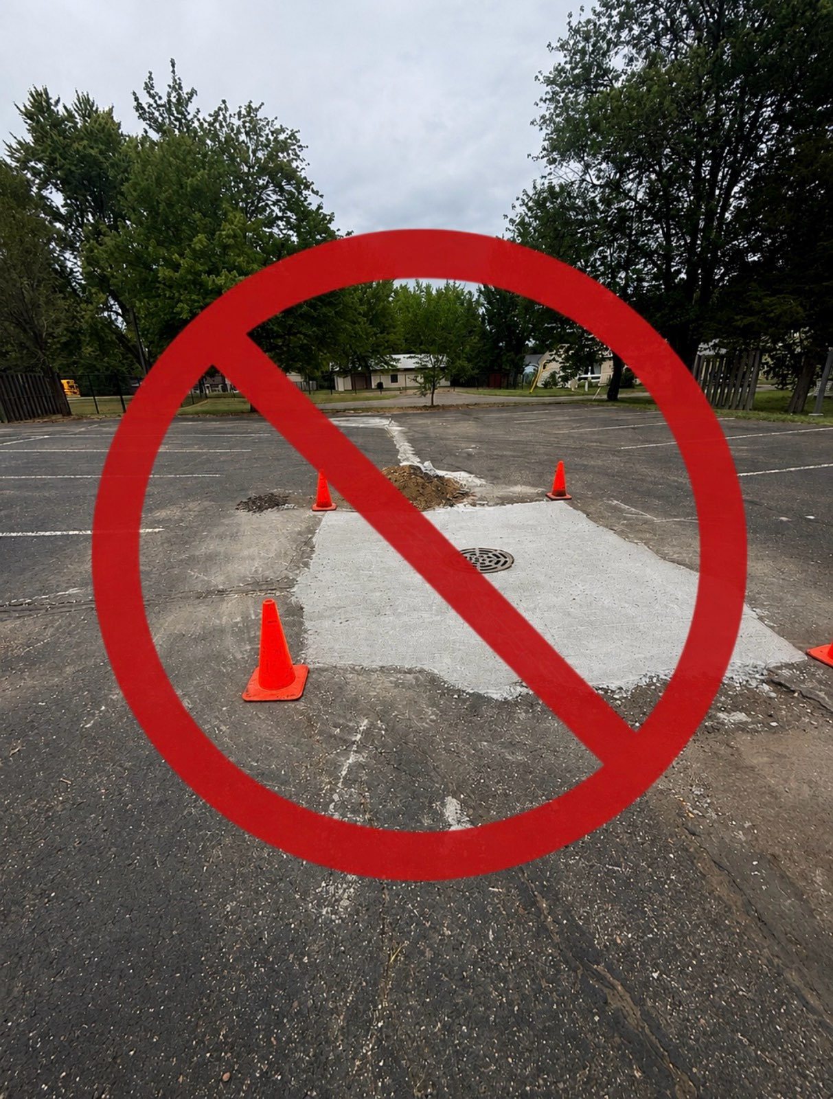 A mismatched concrete patch poured around a storm grate in a cracked school parking lot, marked with a red no symbol